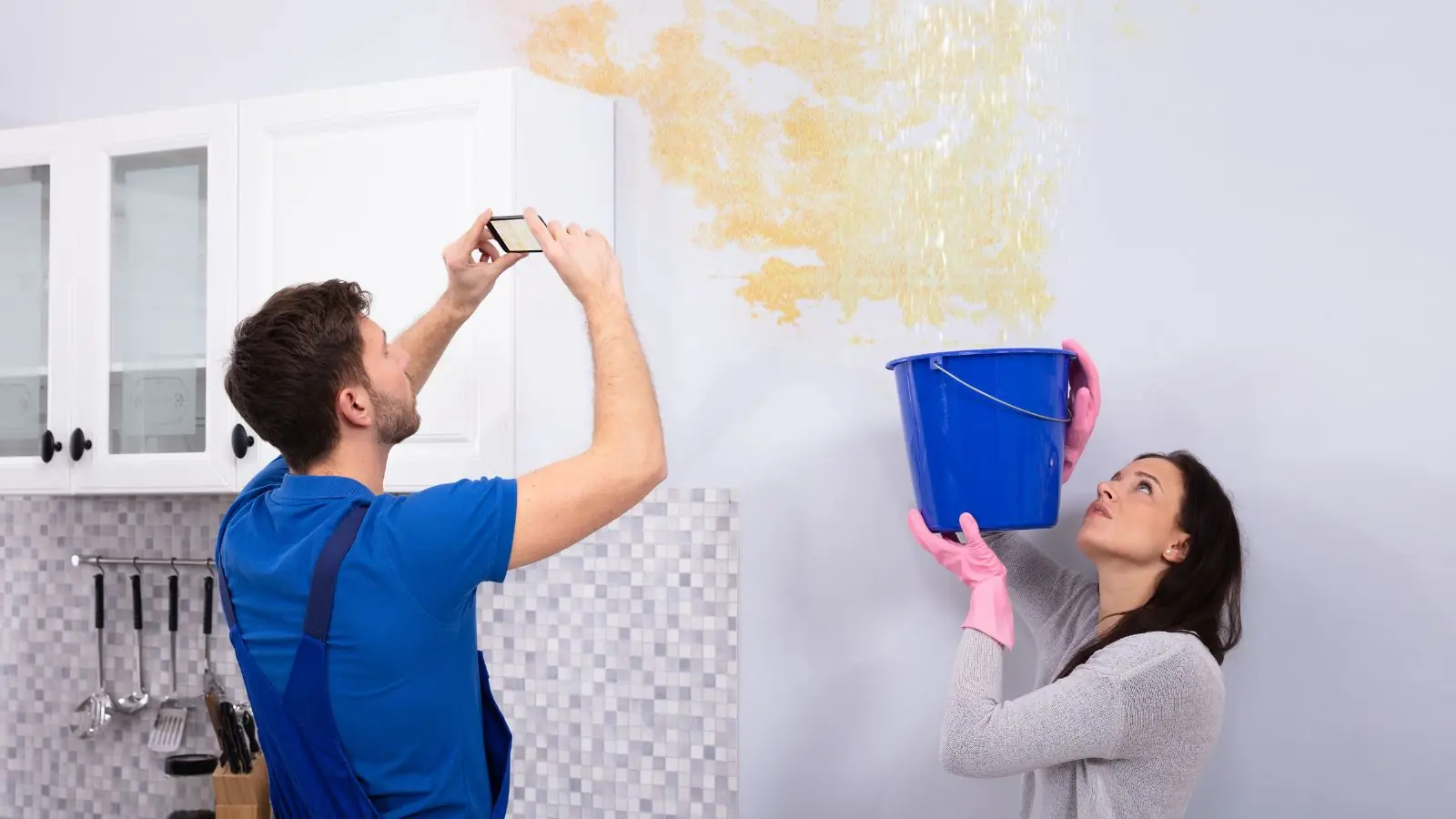 Man photographing a stained wall while a woman holds a bucket under a ceiling leak