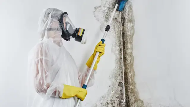 Technician in a respirator and protective suit working on a wall with a long-handled tool