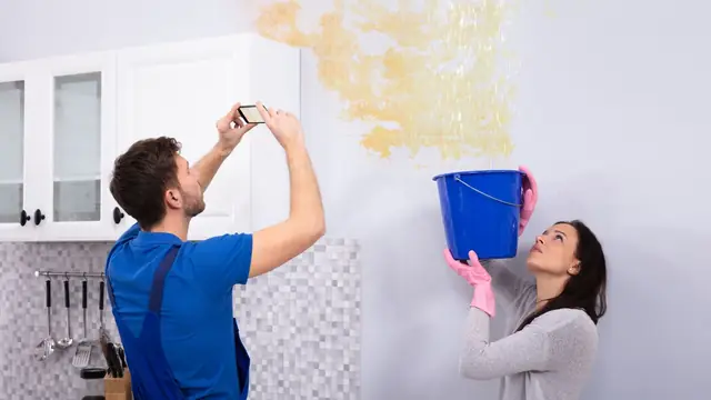 Man photographing a stained wall while a woman holds a bucket under a ceiling leak